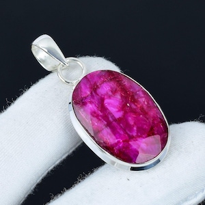 May include: A vibrant, oval-shaped pendant featuring a large, faceted ruby gemstone. The ruby has a rich, pinkish-red hue and is set in a polished silver bezel. The pendant includes a silver bail for attaching to a chain.