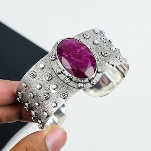 May include: A silver cuff bracelet with a large, oval-shaped, deep pink gemstone set in a decorative silver bezel. The bracelet has a textured surface with small, raised dots.