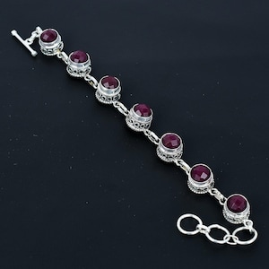May include: A silver bracelet featuring six round, deep red gemstones. The gemstones are set in ornate, detailed silver settings and connected by a delicate chain. The bracelet has a toggle clasp closure.