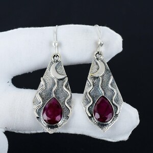 Peut inclure: Une paire de boucles d'oreilles pendantes en argent avec une pierre pr&eacute;cieuse rouge en forme de larme et un charme de croissant de lune. Les boucles d'oreilles ont un design textur&eacute; et ondul&eacute;.