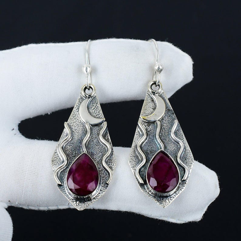 Peut inclure: Une paire de boucles d'oreilles pendantes en argent avec une pierre pr&eacute;cieuse rouge en forme de larme et un charme de croissant de lune. Les boucles d'oreilles ont un design textur&eacute; et ondul&eacute;.