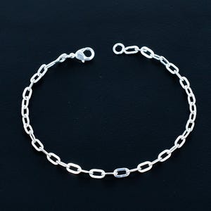 Puede incluir: Una pulsera de cadena plateada con eslabones ovalados. La pulsera tiene un cierre de mosquetón y una pequeña extensión de cadena. La joya se muestra sobre un fondo negro.