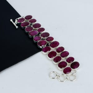 May include: A silver bracelet featuring three rows of oval-shaped, deep red gemstones. The gemstones are set in individual silver bezels and connected by silver links. The bracelet has a toggle clasp for closure.