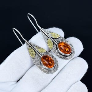 Op de afbeelding: Een paar zilveren oorhangers met ovale stenen in barnsteenkleur. De oorbellen hebben een bladvorm met goudkleurige accenten. De achtergrond is zwart.