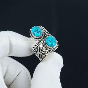May include: A silver-colored ring featuring two oval turquoise stones. The ring has an open design with intricate scrollwork and rope-like detailing around the stones. The turquoise stones are a vibrant blue-green color.