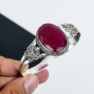 以下が含まれることがあります： 大きな楕円形の赤い宝石と蝶のアクセントが付いたシルバーのバングルブレスレットです。