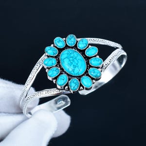 Puede incluir: Brazalete plateado con una piedra turquesa ovalada central rodeada de piedras turquesas más pequeñas. El brazalete tiene un diseño abierto con detalles grabados decorativos a lo largo de la banda. Joyería de turquesa.