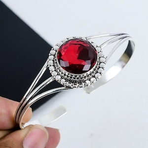 May include: A silver bangle bracelet with a red gemstone set in a silver bezel. The bezel is decorated with small silver beads.