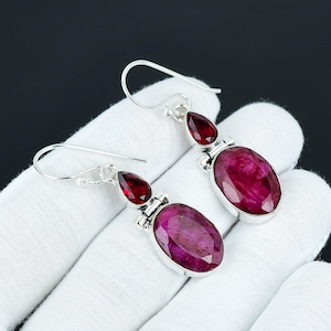 Könnte beinhalten: Ein Paar silberne Ohrringe mit roten Edelstein-Akzenten. Die Ohrringe haben einen tropfenförmigen Edelstein und einen größeren ovalen Edelstein.