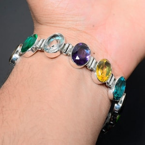May include: A silver bracelet with seven oval-shaped gemstones. The gemstones are green, blue, white, purple, yellow, blue, and green.