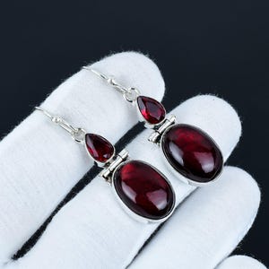 Op de afbeelding: Een paar zilveren oorhangers met rode edelsteen druppels. De oorbellen hebben een traanvormige edelsteen aan de bovenkant en een grotere ovale edelsteen aan de onderkant.