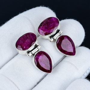 May include: A pair of silver earrings featuring oval and pear-shaped, faceted, deep red gemstones. The earrings are set in a silver metal with a decorative clasp design. The gemstones have a rich, saturated color and are displayed on a white surface.