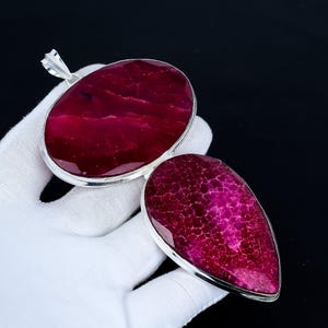 Pode incluir: Dois pingentes com uma pedra vermelha escura e texturizada em metal prateado. Um pingente é oval, o outro em forma de gota. Os pingentes são exibidos em um fundo preto.