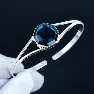 Pode incluir: Uma pulseira de punho prateada com uma grande pedra preciosa azul escura facetada e octogonal no centro. A pedra preciosa é engastada num design minimalista e aberto. A pulseira é aberta para facilitar o uso.