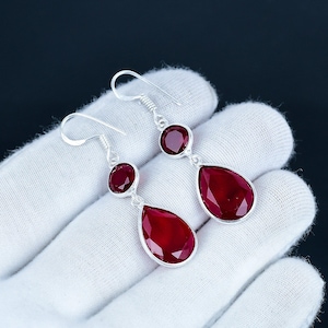 May include: A pair of silver dangle earrings with three red gemstones each. The top gemstone is round, the middle is round, and the bottom is a teardrop shape. The earrings are displayed on a white surface.