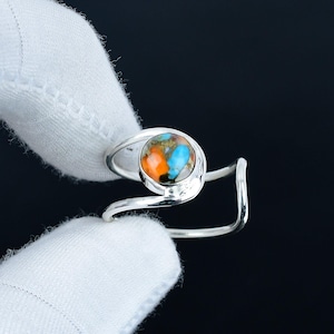 Puede incluir: Anillo de plata con una piedra redonda multicolor con tonos naranja, azul y verde. El anillo tiene un diseño abierto único con una banda curva. La piedra está engastada en un bisel. Fondo oscuro.