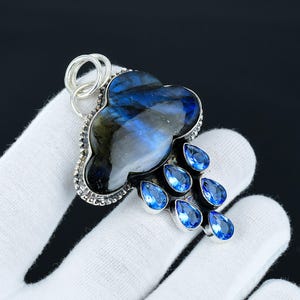 May include: A silver pendant featuring a dark blue labradorite gemstone carved into the shape of a cloud with a silver border.  The cloud is adorned with seven blue topaz gemstones, resembling raindrops.