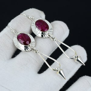 May include: A pair of silver dangle earrings featuring red ruby gemstones. The earrings have a round, bezel-set ruby stone surrounded by a silver frame. The earrings hang from a silver chain with a pointed tip.