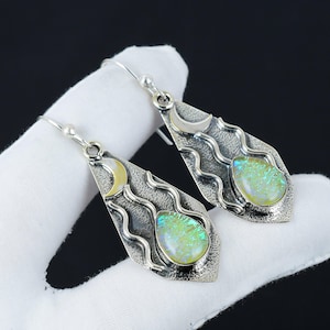 Può includere: Un paio di orecchini pendenti color argento. Ogni orecchino presenta una pietra di opale iridescente a forma di goccia, incastonata in una cornice strutturata. Una luna crescente e linee ondulate sono anch'esse incorporate nel design. Gli orecchini sono mostrati su una superficie bianca.