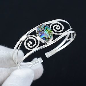 Peut inclure: Un bracelet manchette en argent avec une pièce maîtresse ovale en coquille d'ormeau. La coquille présente des reflets irisés bleus, verts et violets. Le bracelet comporte des motifs tourbillonnants décoratifs et est ouvert en bas.