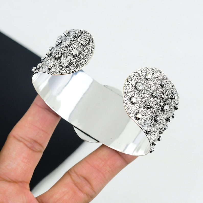 May include: A silver cuff bracelet with a textured surface and small raised dots. The bracelet is open and has a curved shape.