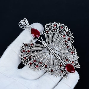 May include: A silver butterfly pendant with detailed filigree work. The pendant is adorned with small, round, red gemstones and two larger, teardrop-shaped red gemstones. The pendant is displayed against a black backdrop.