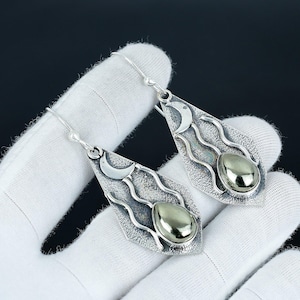 May include: Silver-tone dangle earrings with a crescent moon and wavy design. Each earring features a teardrop-shaped pyrite stone. The earrings are displayed against a dark background.