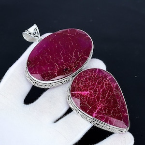 May include: Two large, oval and teardrop-shaped pendants with a deep red gemstone set in ornate silver-coloured metal. The gemstones have a cracked appearance. The pendants are displayed on a white surface.