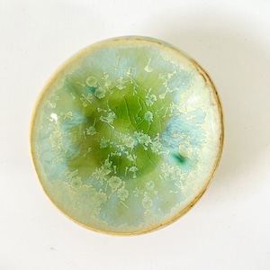 May include: A small, round ceramic dish with a green and blue glaze. The glaze has a unique, speckled pattern.
