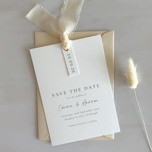 Puede incluir: Una invitación de boda color crema con el texto "Save the Date" y los nombres "Emma & Aaron". La invitación está atada con una cinta crema e incluye una etiqueta con la fecha "26.09.26". Un tallo de flor seca está a la derecha.