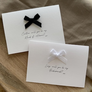 May include: Two white cards with black and white bows. The top card has a black bow and the text "Carttia, will you be my Maid of Honour?" The bottom card has a white bow and the text "Lucy, will you be my Bridesmaid?"