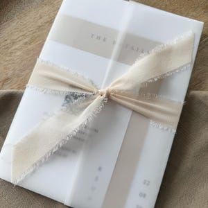 May include: A stack of white cards wrapped in translucent paper and tied with a cream-colored ribbon. The ribbon has frayed edges and is knotted in the center. The text "THE DETAILS" is visible on the top card.