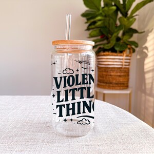 Puede incluir: Una botella de agua de vidrio transparente con tapa de bamb&uacute; y una pajita. La botella tiene un gr&aacute;fico negro que dice "VIOLENT LITTLE THING" con estrellas y nubes.