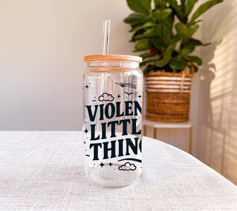Puede incluir: Una botella de agua de vidrio transparente con tapa de bamb&uacute; y una pajita. La botella tiene un gr&aacute;fico negro que dice "VIOLENT LITTLE THING" con estrellas y nubes.
