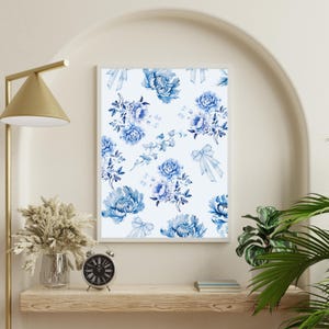 May include: A framed floral print featuring a repeating pattern of blue peonies and bows on a white background. The artwork is displayed on a wooden shelf with a gold lamp, a vase of dried flowers, and a small clock.
