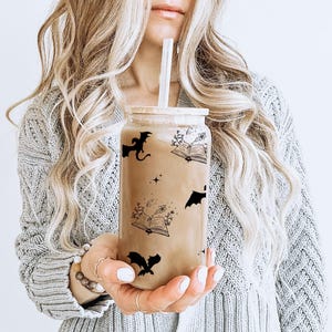 Puede incluir: Un vaso de vidrio transparente con un dise&ntilde;o en blanco y negro con murci&eacute;lagos, libros y estrellas. El vaso est&aacute; lleno de un l&iacute;quido marr&oacute;n y tiene una pajita.