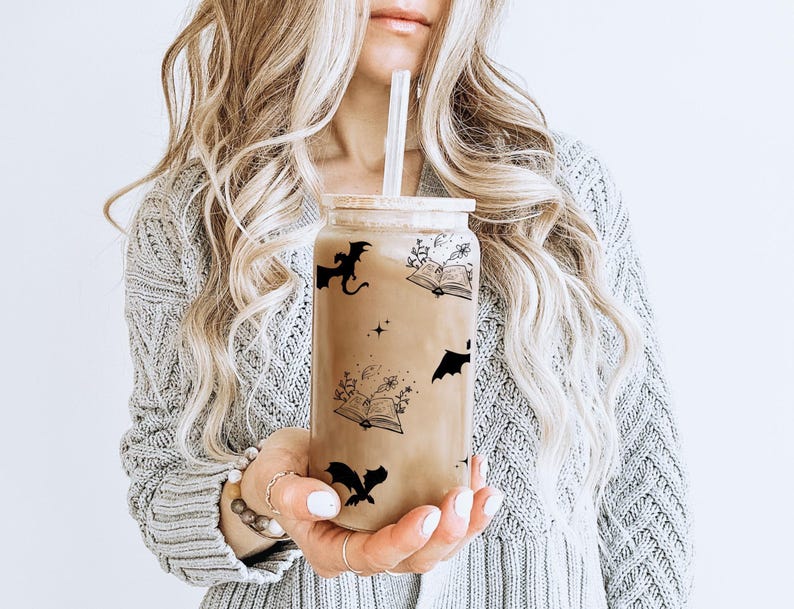 Puede incluir: Un vaso de vidrio transparente con un dise&ntilde;o en blanco y negro con murci&eacute;lagos, libros y estrellas. El vaso est&aacute; lleno de un l&iacute;quido marr&oacute;n y tiene una pajita.