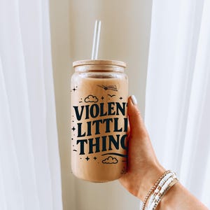 Puede incluir: Un tarro de vidrio con tapa marr&oacute;n y un gr&aacute;fico en blanco y negro que dice "VIOLENT LITTLE THING" con nubes y estrellas. El tarro est&aacute; lleno de un l&iacute;quido marr&oacute;n claro y tiene una pajita que sobresale de la parte superior.