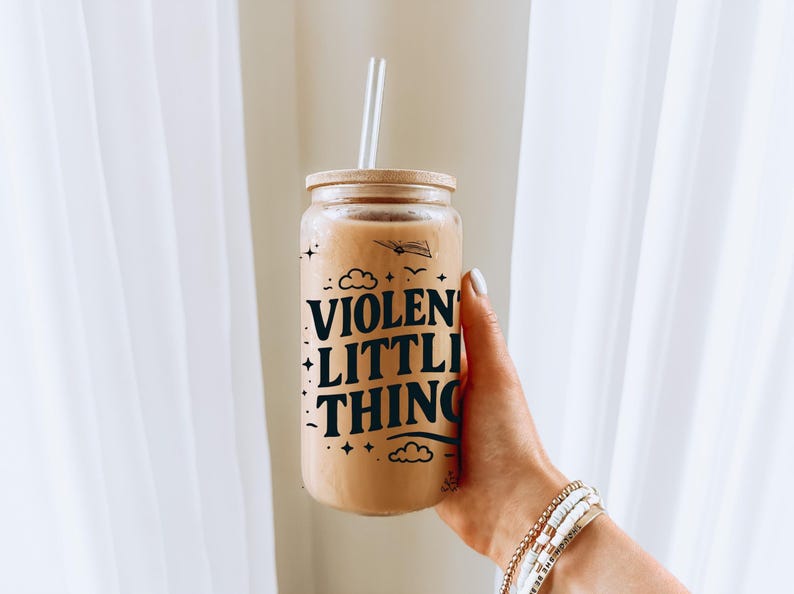 Puede incluir: Un tarro de vidrio con tapa marr&oacute;n y un gr&aacute;fico en blanco y negro que dice "VIOLENT LITTLE THING" con nubes y estrellas. El tarro est&aacute; lleno de un l&iacute;quido marr&oacute;n claro y tiene una pajita que sobresale de la parte superior.