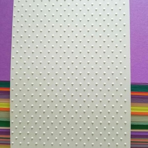 May include: A rectangular white cardstock sheet featuring a uniform, embossed pattern of small, raised dots arranged in a grid. The surface texture is consistent across the entire piece, creating a tactile, repeating polka dot design.
