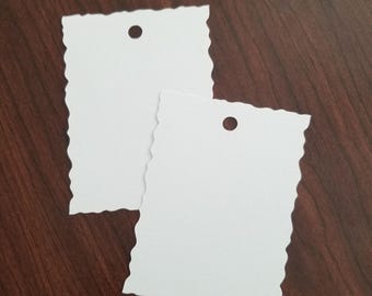 May include: Two white rectangular tags with a decorative, wavy edge. Each tag has a small, circular hole at the top. The tags are blank and ready for customization. The tags are on a dark brown wooden surface.