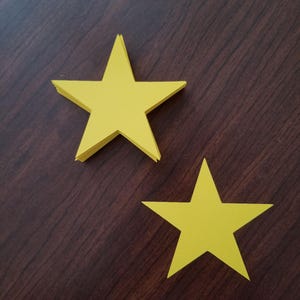 May include: Two yellow paper stars on a dark brown wooden surface. The larger star is on top of the smaller star.