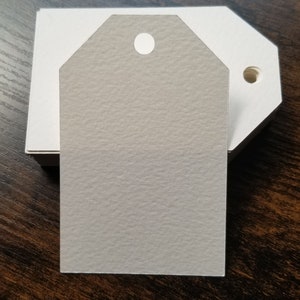 May include: A blank gray gift tag with a round hole at the top. The tag is sitting on a stack of white card stock.