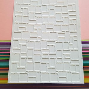 May include: A rectangular white embossing folder featuring a raised, textured pattern of irregular, interlocking geometric blocks. The surface has a matte finish with deep indentations that create a three-dimensional grid effect across the entire sheet.