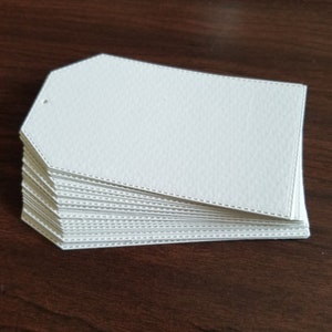 May include: A stack of rectangular white paper tags with angled top corners. Each tag features a textured surface and a decorative stitched border pattern embossed along the edges. A small circular hole is punched near the top left corner.