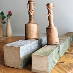 May include: Two wooden mallets with leather straps, resting on a wooden surface. The mallets have a dark brown stain and a black band around the top.
