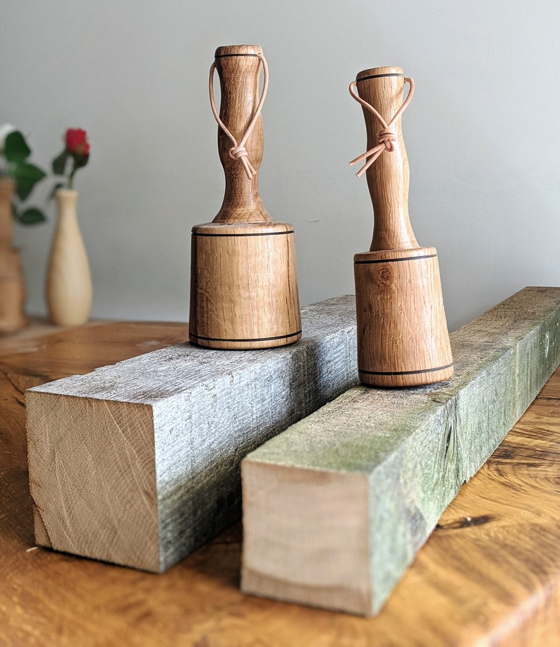 May include: Two wooden mallets with leather straps, resting on a wooden surface. The mallets have a dark brown stain and a black band around the top.