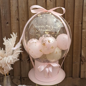 May include: A pink gift box topped with a clear balloon containing smaller pink and white balloons. The clear balloon has gold confetti and the text "Congrats On Your Masters". A pink ribbon adorns the box and balloon.