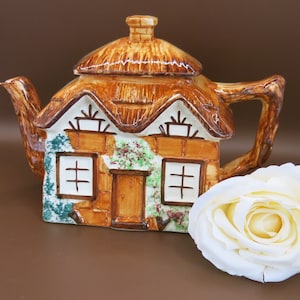 May include: A brown and white ceramic teapot shaped like a cottage with a thatched roof. The teapot has a spout and handle and is decorated with green and brown accents.
