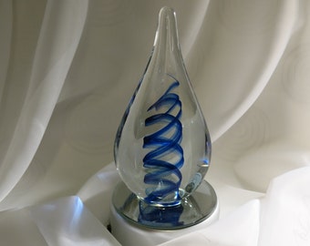 May include: A clear glass teardrop-shaped sculpture with a blue spiral design inside. The sculpture sits on a small, round, mirrored base, and is set against a white fabric background.